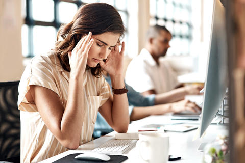 Junge Frau sitzt im Büro am Computer und greift sich mit geschlossenen Augen gestresst an die Schläfen.