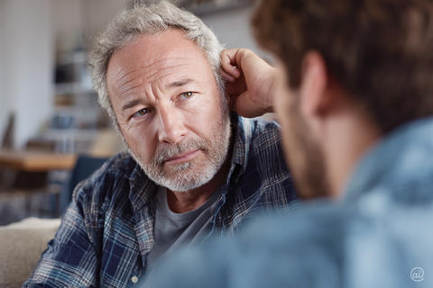 Mann mittleren Alters mit grauem Bart signalisiert Hörprobleme und spricht mit seinem Sohn.