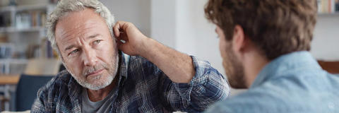 Mann mittleren Alters mit grauem Bart signalisiert Hörprobleme und spricht mit seinem Sohn.
