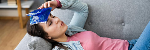 Junge Frau liegt mit geschlossenen Augen auf einer Couch und nutzt ein blaues Kühlpad auf der Stirn, um Symptome wie Kopfschmerzen zu behandeln.