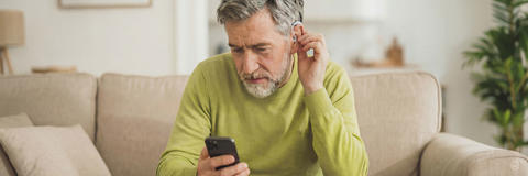 Ein älterer Mann sitzt auf einem Sofa, sieht konzentriert auf sein Smartphone und passt dabei ein Hörgerät an seinem Ohr an.