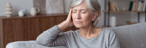 Ältere Frau mit grauen Haaren sitzt mit geschlossenen Augen und hält sich den Kopf wegen Ohrgeräuschen