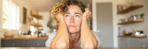 Psychische Belastung durch Tinnitus: Eine Frau sitzt sichtlich erschöpft und überfordert am Küchentisch, den Kopf in die Hände gestützt.