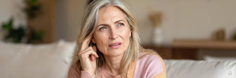 Frau mit langen, grauen Haaren blickt nachdenklich und berührt ihr Ohr, möglicherweise Anzeichen für Ohrbeschwerden.