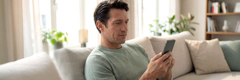 Entspannt sitzender Mann nutzt sein Smartphone auf dem Sofa im Wohnzimmer.