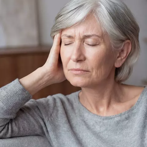 Ältere Frau mit grauen Haaren sitzt mit geschlossenen Augen und hält sich den Kopf wegen Ohrgeräuschen