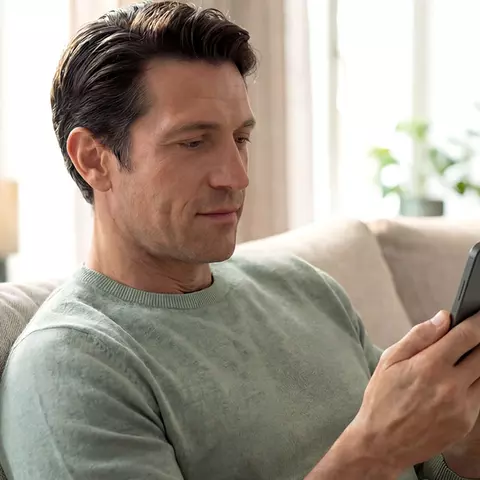 Entspannt sitzender Mann nutzt sein Smartphone auf dem Sofa im Wohnzimmer.